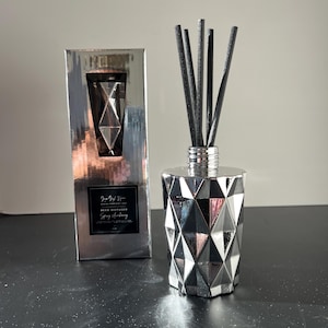 Puede incluir: Un difusor de caña plateado con un diseño geométrico y cañas negras. La caja de al lado tiene el mismo diseño y el texto "Just Girl Home" y "Reed Diffuser Spring Ambience".