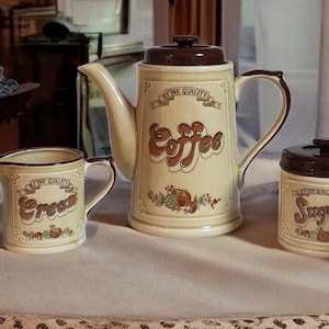 May include: A vintage coffee set with a cream-colored finish and brown accents. The set includes a coffee pot, two mugs, a creamer, and a sugar bowl. Each piece is decorated with the word "Coffee" or "Sugar" in a retro font.