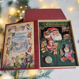 May include: A vintage Christmas-themed book with a red cover and a decorative interior. The left side features a musical score with cherubs and instruments. The right side has a Santa Claus figure and other festive characters, with the word "Christmas" at the top.