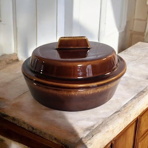 May include: A brown ceramic casserole dish with a lid. The dish has a rounded shape with a slightly flared rim. The lid features a rectangular handle. The dish is glazed with a glossy finish, and the color is a rich, dark brown.
