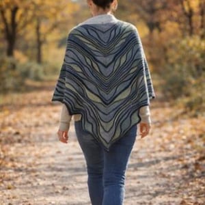 May include: A triangular knit shawl featuring a wavy, chevron-like pattern in shades of navy blue, sage green, and cream. The shawl is worn over a light-colored long-sleeved top and blue denim jeans, displaying a textured, multi-colored yarn design.