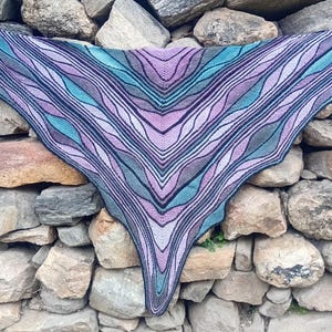 May include: A triangular knit shawl featuring a repeating wave pattern in shades of purple, lavender, teal, and dark gray. The garment is constructed with a central spine and symmetrical color-blocked sections that create a fluid, rippled visual effect.