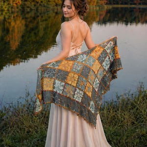 May include: A triangular crochet shawl featuring a patchwork pattern of square motifs in shades of mustard yellow, sage green, and charcoal gray. The shawl has a delicate lacy texture with small decorative bobbles along the outer edges.