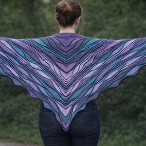 May include: A triangular knit shawl featuring a wavy, multi-colored pattern in shades of purple, lavender, teal, and dark blue. The garment has a scalloped edge and a symmetrical design that radiates from the center point down to the bottom hem.