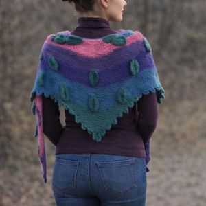 May include: A triangular crochet shawl featuring horizontal stripes in shades of pink, purple, blue, and teal. The shawl is decorated with small, raised, leaf-shaped appliques in a dark teal color and has a scalloped edge along the bottom border.