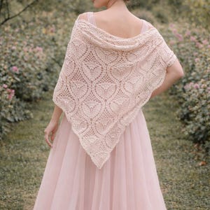 May include: A pale pink shawl with an openwork pattern is worn over the shoulders. The shawl's detailed design is set against a pink dress and a garden background. The shawl appears to be made of a soft, lightweight material, suitable for a special occasion.