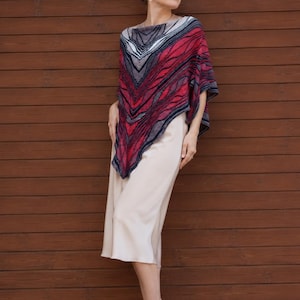 May include: A knitted poncho featuring a vibrant red, black, and white abstract wave pattern with a pointed hemline, worn over a solid cream-colored midi skirt and light-colored strappy high-heeled sandals. The poncho has a wide neckline.