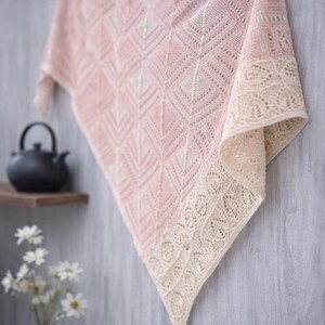 May include: A delicate, hand-knitted shawl in soft pink and cream. The shawl features a geometric pattern in the center and a decorative lace border. The shawl is displayed against a gray background, with a teapot and flowers in the background.