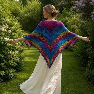 May include: A woman wearing a colorful, rainbow-patterned shawl over a white dress, walking on a green grassy path. The shawl features a V-neck design with shades of blue, purple, yellow, and pink. The background includes lush green foliage and trees.
