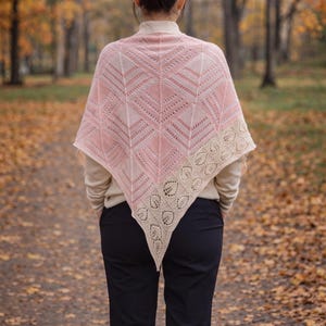 May include: A pink and cream-colored shawl with a geometric pattern and a decorative border. The shawl is triangular and appears to be knitted or crocheted. The person is wearing a cream-colored sweater and navy blue pants.
