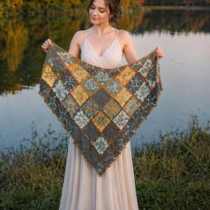 May include: A triangular crochet shawl featuring a geometric pattern of diamond-shaped motifs in shades of mustard yellow, dark gray, and muted teal. The edges are finished with a decorative picot border, and the piece is constructed from yarn.