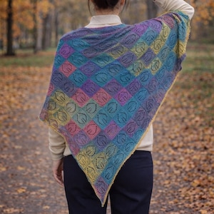 May include: A triangular knit shawl featuring a multicolored gradient pattern of blue, purple, pink, and yellow squares. Each square contains a delicate leaf-shaped lace cutout design. The shawl is draped over a beige long-sleeve shirt and black trousers.