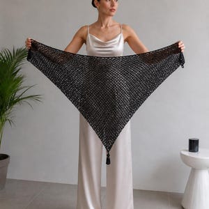 May include: A black triangular crochet shawl featuring an openwork mesh pattern and a single decorative tassel at the bottom point. The accessory is crafted from dark yarn and displays a consistent geometric stitch throughout its wide span.
