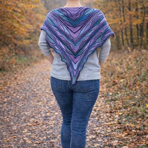 May include: A triangular knit shawl featuring a gradient of purple, violet, and blue tones with a textured, wavy striped pattern. The shawl is draped over a light gray long-sleeve shirt and blue denim jeans, showcasing a pointed hemline.