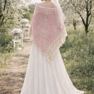 May include: A triangular knit shawl featuring a soft pink center with a geometric chevron pattern and a wide, cream-colored lace border with an intricate leaf motif. The shawl is draped over the shoulders of a long, flowing white gown.