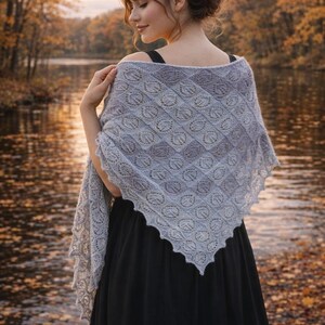 May include: A light gray triangular shawl featuring an intricate knitted lace pattern with repeating leaf motifs and a scalloped edge. The shawl is draped over the shoulders, showcasing the delicate openwork texture and the soft yarn construction.