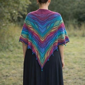 May include: A triangular knit shawl featuring a vibrant, multi-colored gradient pattern of wavy stripes in shades of magenta, purple, blue, and teal. The shawl has a pointed hemline adorned with small, dangling tassels along the bottom edge.