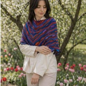 May include: A triangular knit shawl featuring a vibrant, wavy pattern in shades of deep blue, bright red, and purple. The shawl is draped over a light beige long-sleeved button-down shirt and matching light beige trousers, creating a layered look.