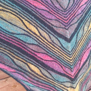 May include: A triangular knitted shawl featuring a vibrant, multi-colored pattern of wavy stripes in shades of magenta, teal, and mustard yellow, separated by bold black yarn lines that create a distinct geometric texture throughout the fabric.
