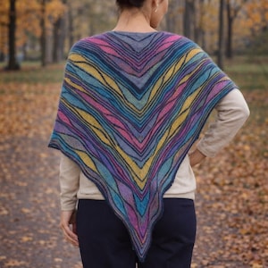 Pode incluir: Um xale listrado multicolorido em tons de azul, roxo, amarelo e rosa. O xale é drapeado sobre os ombros, criando uma forma em V. O fundo apresenta um caminho com folhas caídas e árvores.