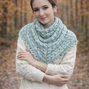 Puede incluir: Un chal triangular de ganchillo en color verde menta claro con un intrincado patrón de encaje y borde festoneado, usado sobre un suéter de punto trenzado color crema y combinado con una diadema estampada en tonos naranja, marrón y amarillo.