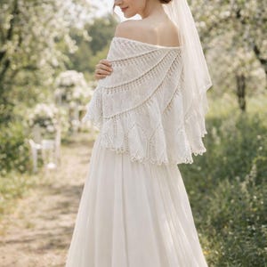 May include: A white off-the-shoulder knitted shawl with a textured lace pattern and scalloped edges, worn over a floor-length white dress with a sheer veil attached to the hair, featuring a flowing skirt and a delicate knit construction.