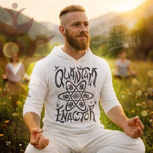 Op de afbeelding: Een witte hoodie met de zwarte woorden "Quantum Enactor", met een gestileerd atoomontwerp. De persoon zit in kleermakerszit in een veld, met een vage achtergrond van andere mensen en de natuur. De hoodie heeft lange mouwen.