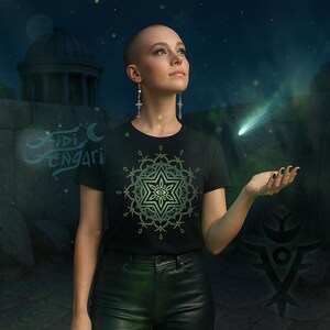 May include: Black t-shirt with a green and white geometric design featuring an eye. The shirt is worn by a person with short hair, wearing earrings, and black trousers. A comet is in the background.