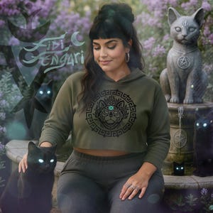 May include: An olive green cropped hoodie with a black cat graphic and the text "Fidi Sengari". The image features a woman with black cats and cat statues. The cats have glowing eyes. The background has purple flowers.