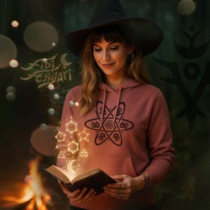 Puede incluir: Una mujer con un sombrero de bruja negro y una sudadera con capucha rosa polvoriento con un diseño gráfico negro. Sostiene un libro abierto con formas geométricas brillantes y el texto "Fibi Fengari". El fondo es un bosque oscuro.