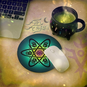 May include: A teal and black round mousepad with a star-like design featuring hands and an eye in the center. A white wireless mouse sits on the pad. A black cauldron mug with green liquid and a laptop are also in the image.