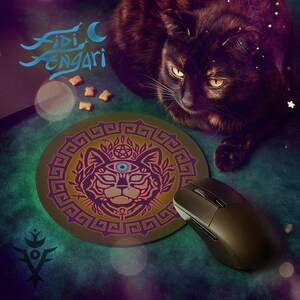 Op de afbeelding: Een ronde muismat met een kattenontwerp in paars en goud, naast een zwarte computermuis. De muismat heeft een kattengezicht met een derde oog, omgeven door een geometrische rand. De tekst "Fidi Engari" staat linksboven.