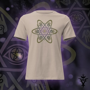 May include: A beige t-shirt featuring a geometric design with hands, an eye, and star shapes in olive green and purple. The design is centered on the front of the short-sleeved shirt. The shirt is displayed on a dark purple background.