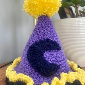 May include: Purple crocheted party hat with a yellow pom-pom on top and a blue felt letter C on the front. The hat has a yellow and blue ruffled trim.