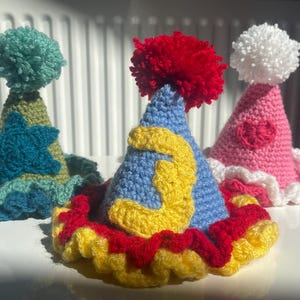 May include: Three crocheted party hats with pom-poms. The hats are blue, green, and pink with yellow, red, and white accents.