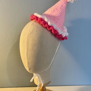 May include: A pink and white crocheted party hat with a white pom-pom on top. The hat is on a white mannequin head.