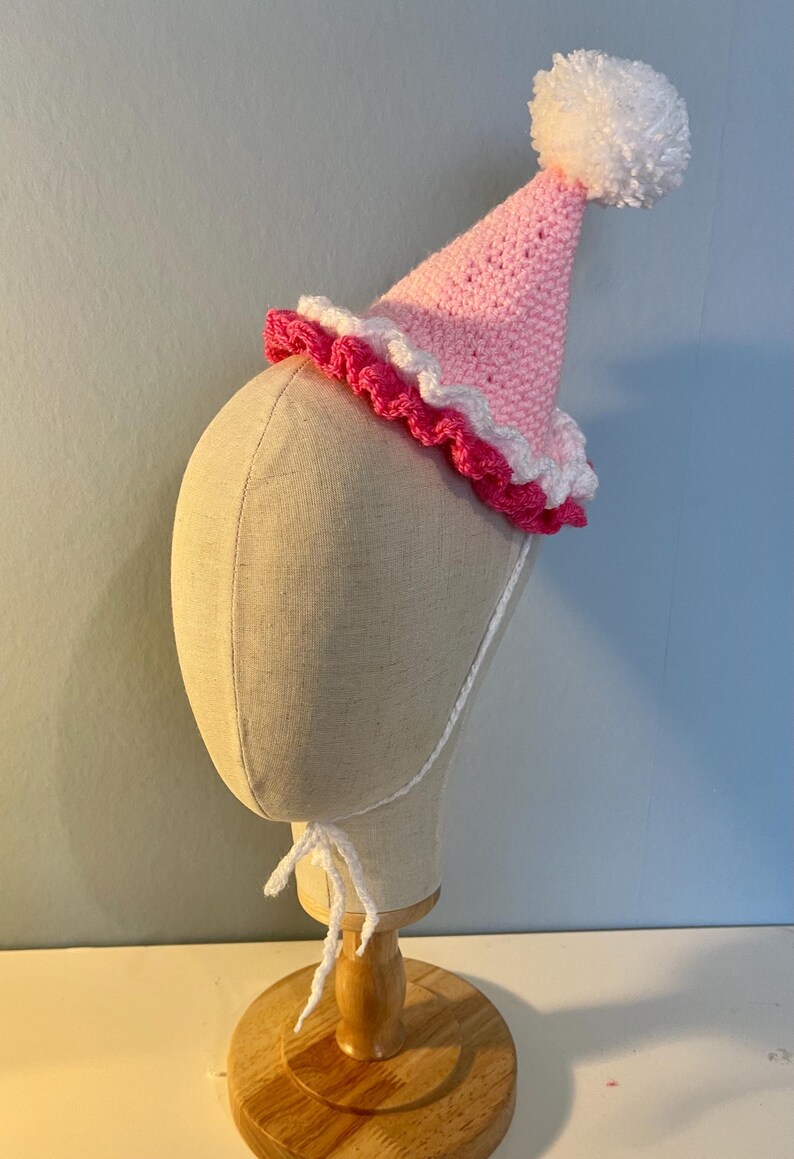 May include: A pink and white crocheted party hat with a white pom-pom on top. The hat is on a white mannequin head.