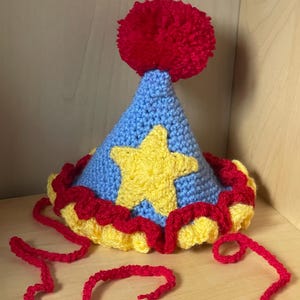 May include: A blue and yellow crocheted party hat with a red pom-pom on top. The hat has a yellow star on the front and a red and yellow ruffle around the bottom.