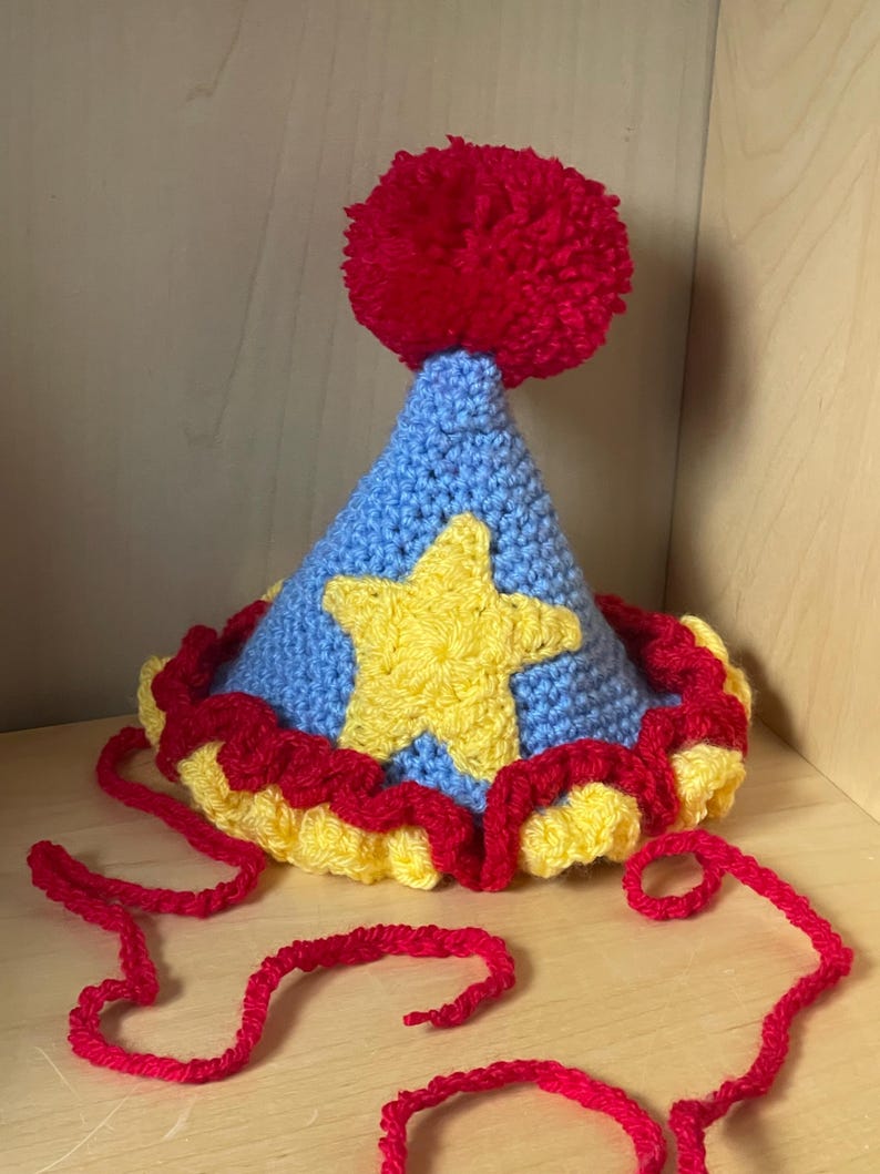 May include: A blue and yellow crocheted party hat with a red pom-pom on top. The hat has a yellow star on the front and a red and yellow ruffle around the bottom.