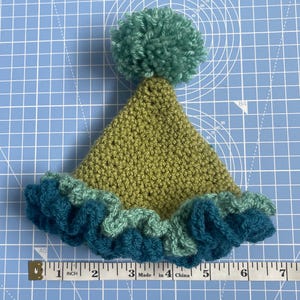 May include: A green crocheted hat with a teal ruffle and a green pom-pom on top.