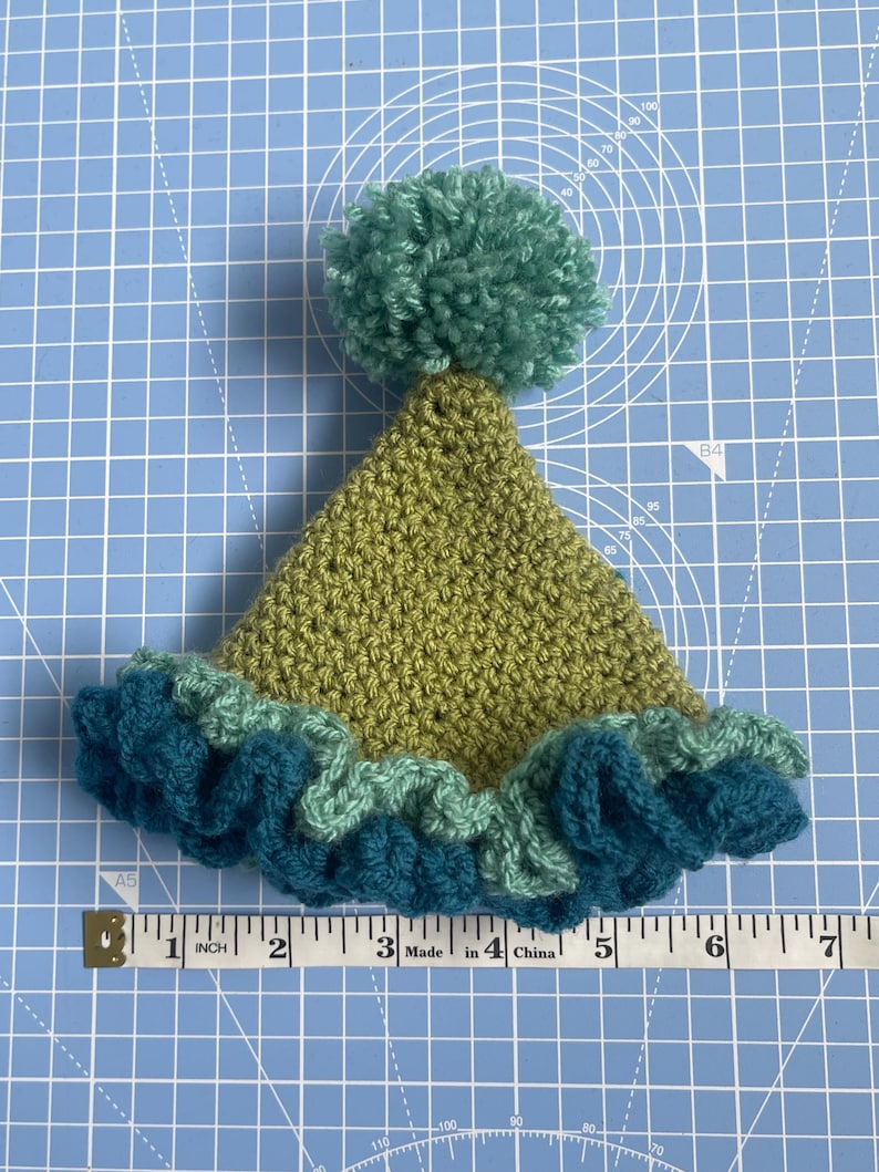 May include: A green crocheted hat with a teal ruffle and a green pom-pom on top.