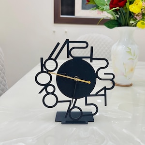 Op de afbeelding: Zwarte metalen klok met gouden wijzers en een rond gezicht. De cijfers 1 tot en met 12 worden weergegeven in een uniek, abstract ontwerp.