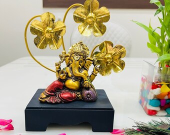 Op de afbeelding: Een gouden Ganesha-beeld met een rode en gouden robe staat op een zwarte basis. Het beeld is omringd door drie gouden bloemvormige decoraties.