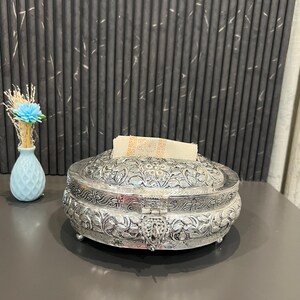 Op de afbeelding: Ovaalvormige, zilverkleurige tissuebox met ingewikkelde bloempatronen. De doos heeft een scharnierend deksel en een decoratieve sluiting. Links staat een kleine blauwe vaas met een blauwe bloem en gedroogde stelen.