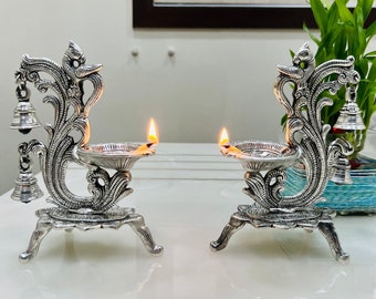 Op de afbeelding: Twee zilveren decoratieve diya-olielampen met sierlijke pauwenmotieven en bellen. De lampen zijn aangestoken met lonten en vlammen.