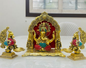 May include: Gold metal statue of Ganesha, the Hindu god of wisdom and prosperity, sitting on a throne with two peacock-shaped oil lamps on either side. The statue is decorated with colorful gemstones.
