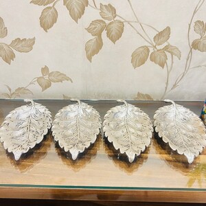 May include: Four silver leaf-shaped serving dishes with intricate designs. The dishes are arranged in a row on a wooden surface.