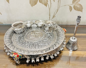 May include: A silver-colored metal plate with intricate designs and a swastika symbol in the center. The plate is surrounded by four small silver bowls and a bell with a handle. The plate is decorated with colorful gemstones.