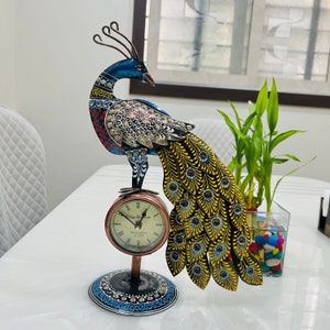 Op de afbeelding: Een decoratieve pauw-klok met een blauwe, groene en gouden afwerking. De klok heeft een rond gezicht met Romeinse cijfers en een zwarte achtergrond. De staartveren van de pauw zijn uitgespreid achter hem.