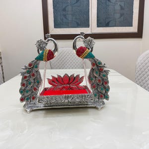 May include: A silver-toned decorative swing featuring two peacock figures with red and teal accents. The base is rectangular with a lotus flower motif, and the central platform is lined with red fabric, suspended by thin silver chains.